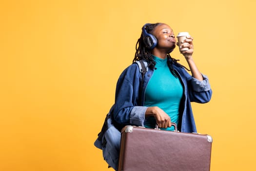 Young girl delighted to go on vacation, travelling with suitcase, drinking coffee, listening music. Woman feeling blissful, enjoying beverage aroma, hearing songs in headphones, studio background