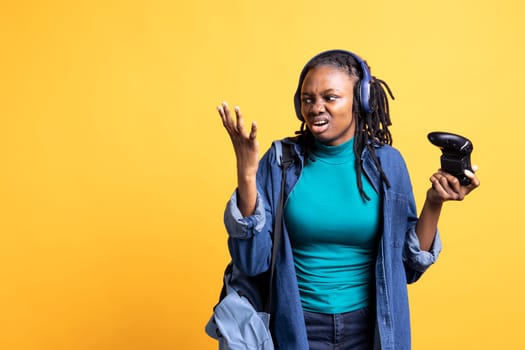 Annoyed BIPOC young girl raging after receiving game over screen, holding controller. Mad african american teenager frustrated after losing videogame, playing with gamepad, studio background