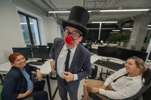 An elderly Caucasian man in a clown costume amuses two Caucasian women in the office