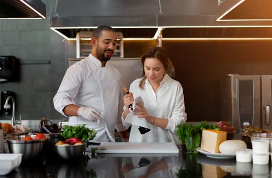 Young diverse male and female chefs in uniform with red wine bottle. Confident professional cooks using wine ingredient at counter in commercial kitchen. Man and woman cooking in professional kitchen.