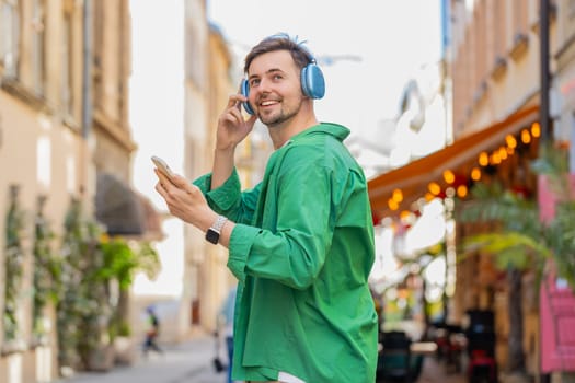 Rear view of happy relaxed man tourist in wireless headphones choosing, listening favorite energetic disco music in smartphone outdoors. Caucasian tourist walking passes by urban sunny city street.