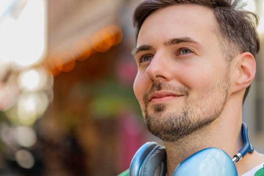 Thoughtful young male tourist in casual clothes with wireless headphones around neck looking up and smiling. Happy positive Caucasian man traveler standing on city urban street. Town lifestyles.
