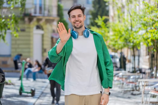 Cheerful young man tourist smiling friendly at camera, waving hands gesturing invitation hello, hi, greeting, goodbye, welcoming with hospitable expression outdoors. Guy on urban city town street.