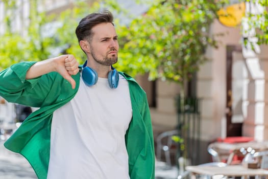 Dislike. Upset Caucasian man showing thumbs down sign gesture, expressing discontent, disapproval, dissatisfied bad work, mistake. Displeased guy male standing on urban city sunshine street outdoors.