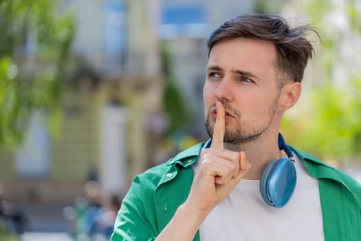 Shh be quiet please. Brunette young man presses index finger to lips makes silence hush gesture sign do not tells gossip secret outdoors. Young guy male stand on urban city street. Town lifestyles.