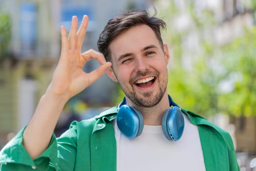Okay. Happy cheerful young Caucasian man looking approvingly at camera showing OK gesture, positive like sign, approve something good, celebrate win. Joyful guy standing in urban city street, outdoors