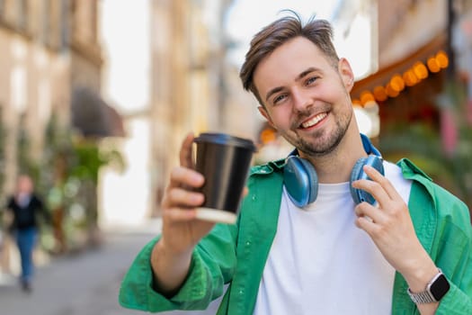 Handsome Caucasian man enjoying morning coffee hot drink and smiling. Relaxing, taking a break. Happy young guy walking on urban city center street, drinking coffee to go. Town lifestyles outside.