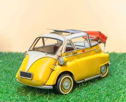 A vintage toy replica depicting a yellow retro microcar with a sunroof and luggage on the rear, parked on a patch of green grass against a sandy yellow background.