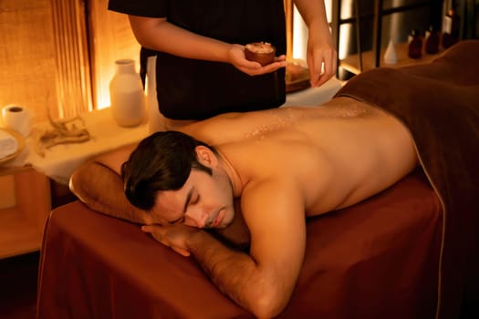 Man customer having exfoliation treatment in luxury spa salon with warmth candle light ambient. Salt scrub beauty treatment in Health spa body scrub. Quiescent