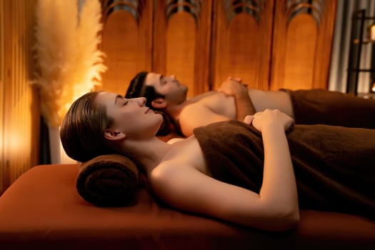 Caucasian couple customer enjoying relaxing anti-stress spa massage and pampering with beauty skin recreation leisure in warm candle lighting ambient salon spa at luxury resort or hotel. Quiescent