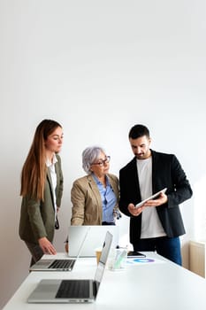 Vertical portrait of group of coworkers working together solving problem looking at laptop and digital tablet in the office. Teamwork and business.