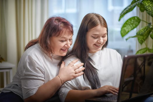 Fat funny funny adult mother and daughter with laptop indoors. A teenage girl teaches a middle-aged woman modern technology. Internet, chatting