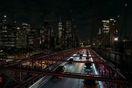 View of cars moving on the Brooklyn Bridge at night with the illuminated skyline of New York City in the background. The photograph captures the busy city life and iconic landmarks.