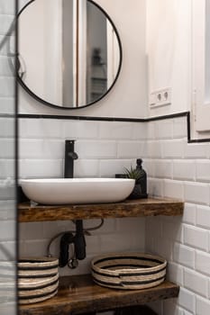 A contemporary white bathroom featuring a round mirror, white tiles, wooden shelves, and woven baskets, creating a stylish and functional space.