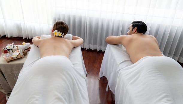 Rear view caucasian couple customer enjoying relaxing anti-stress spa massage and pampering with beauty skin recreation leisure in day light ambient salon spa at luxury resort or hotel. Quiescent