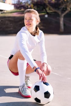 young caucasian woman squatting next to a soccer ball ready to play in a urban football court, concept of sport and active lifestyle