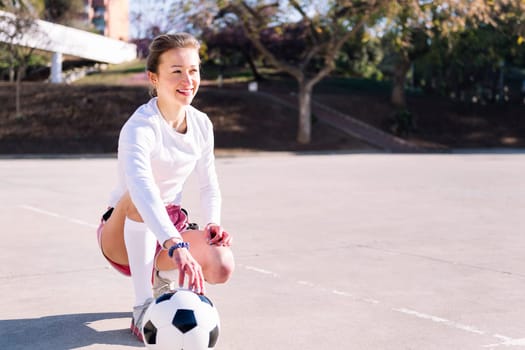 young caucasian woman squatting next to a soccer ball ready to play in a urban football court, concept of sport and active lifestyle, copy space for text