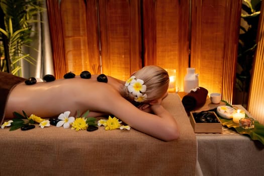 Hot stone massage at spa salon in luxury resort with warm candle light, blissful woman customer enjoying spa basalt stone massage glide over body with soothing warmth. Quiescent