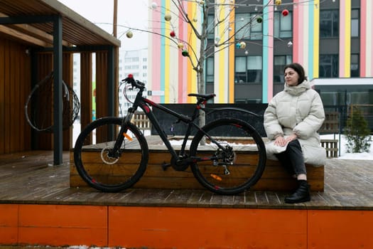 A woman is seated on a bench placed next to a bicycle. She appears relaxed and is looking around. The bike is parked by her side, suggesting she may have just finished a ride.