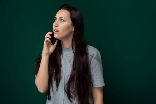 A woman stands in front of a green wall, engaged in a conversation on her cell phone. She appears focused on the call as she gestures with her free hand. The background is a solid, vibrant green color.