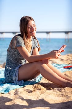 young happy caucasian woman on vacation using smartphone app sitting on towel on the beach
