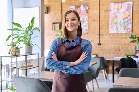 Confident successful young woman service worker owner in apron with crossed arms looking at camera in restaurant cafeteria coffee pastry shop interior. Small business staff occupation entrepreneur work