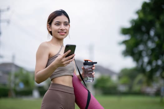 A young woman in athletic wear holding a yoga mat and water bottle, ready for a yoga session in the park, promoting a healthy and active lifestyle.