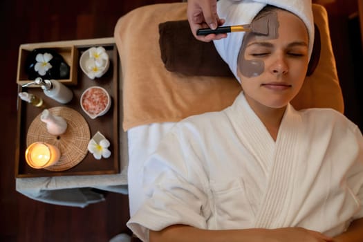 Serene ambiance of spa salon, top view woman customer indulges in rejuvenating with charcoal face cream massage with warm lighting candle. Facial skin treatment and beauty care concept. Quiescent