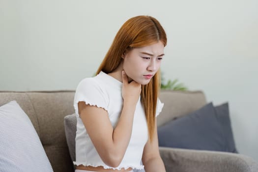 Young woman feeling unwell at home, sitting on a couch and holding her throat, depicting sickness and discomfort in a home environment.