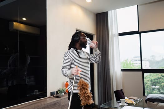 A man cleans a modern home with a feather duster, listening to music and drinking water in a bright, spacious living room.