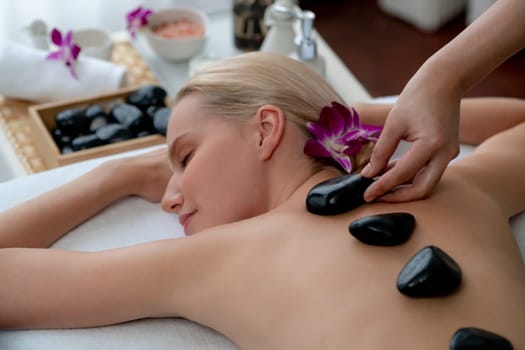 Hot stone massage at spa salon in luxury resort with day light serenity ambient, blissful woman customer enjoying spa basalt stone massage glide over body with soothing warmth. Quiescent