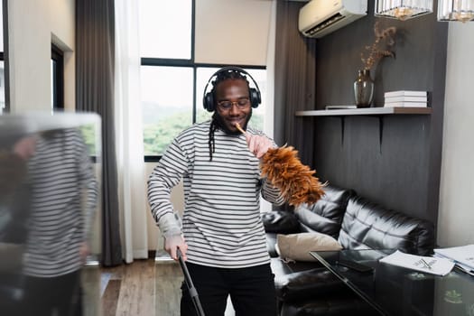 A man wearing headphones cleans a modern living room with a feather duster, enjoying the process.