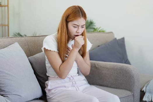 Young woman feeling unwell, coughing and holding chest while sitting on a sofa at home, depicting illness and recovery.
