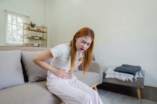 Young woman experiencing stomach pain while sitting on a couch at home, illustrating the concept of being sick and unwell in a modern living room.