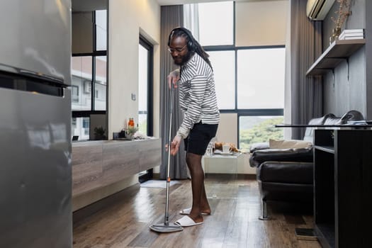 Man mopping floor in modern living room, wearing headphones. Bright, airy space with large windows and contemporary furniture.