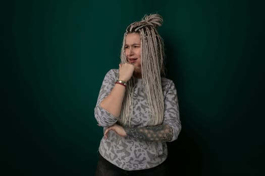 A woman with long blonde dreadlocks is seen standing in front of a vibrant green wall, exuding a confident demeanor. Her hair falls in intricate twists, contrasting against the bold background.