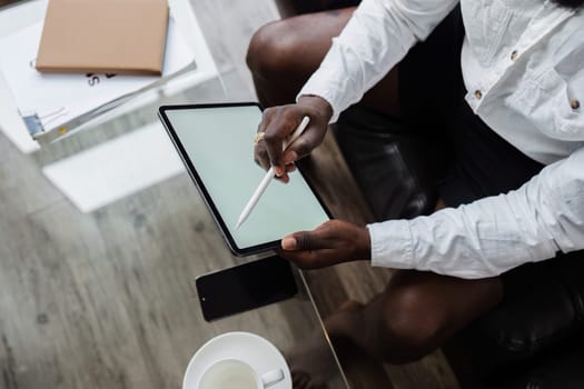 A person working remotely using a tablet and stylus, with a smartphone and coffee on a glass table in a modern home office setting.