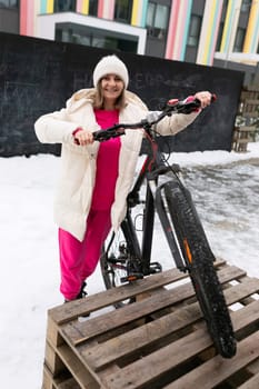 A woman standing next to a bicycle in a snow-covered area. The woman appears to be preparing to ride the bicycle or adjusting something on it. The snow around them is undisturbed, suggesting recent snowfall.