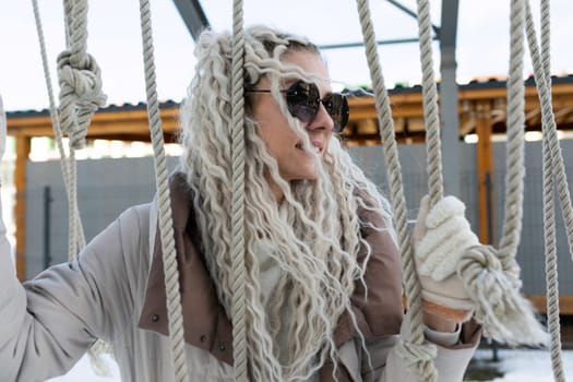 A woman with flowing long white hair is suspended on a rope with a serious expression. She appears to be carefully balancing herself while gripping the rope tightly.