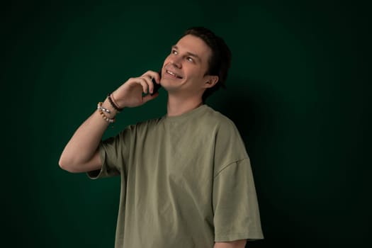 A man in casual clothing stands in front of a bright green wall, engaged in a phone conversation. He holds a cell phone to his ear as he gestures and talks animatedly.