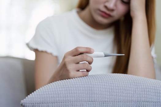 Young woman feeling unwell at home, holding a thermometer, resting on the couch, experiencing fever and fatigue.