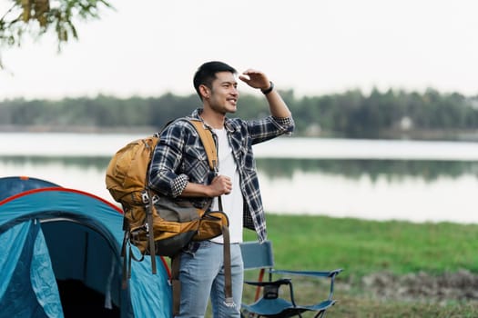 A young man with a backpack standing near a tent by a lake, enjoying the view. Perfect for travel, adventure, and camping themes.
