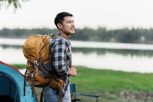 Young traveler with a backpack standing by a lake, surrounded by nature, and a tent in the background. Perfect for travel and camping concepts.