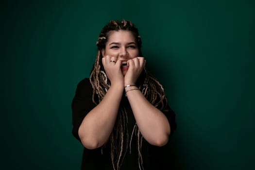 A woman with dreadlocks is standing in front of a solid green wall. She is looking straight ahead, with a calm expression on her face. The sunlight casts a soft shadow behind her, highlighting her features.