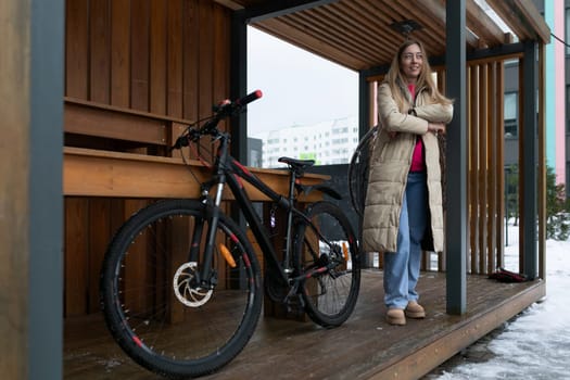 A woman standing on a porch next to a bike, with a relaxed expression on her face. The porch is wooden and the bike is leaning against a railing.