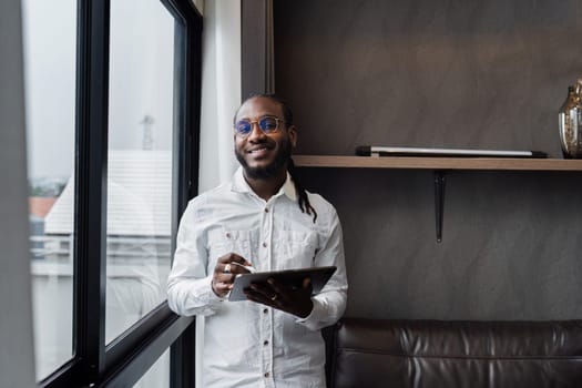 A professional man working remotely from home, using a tablet while standing by a window in a stylish home office.