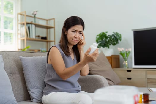 Middle-aged woman carefully reading a medication bottle label while sitting on a comfortable couch in a well-lit, modern living room.