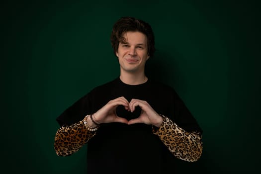 A man is standing, forming a heart shape with his hands in front of his chest. The background is blurred, emphasizing the gesture.