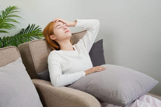 Young woman feeling unwell at home, resting on a sofa with a hand on her forehead, experiencing sickness or fatigue in a cozy living room setting.