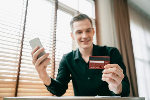 Young man sitting at table using online payment app and digital wallet on smartphone to pay with credit card. E commerce shopping and modern purchasing via mobile internet. Unveiling